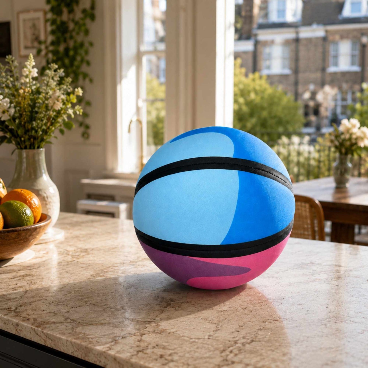 A colorful HouseHoops basketball - SH  with a light blue top half, a pink bottom half, and two black horizontal bands, sitting on a marble kitchen counter in a bright, sunlit kitchen.