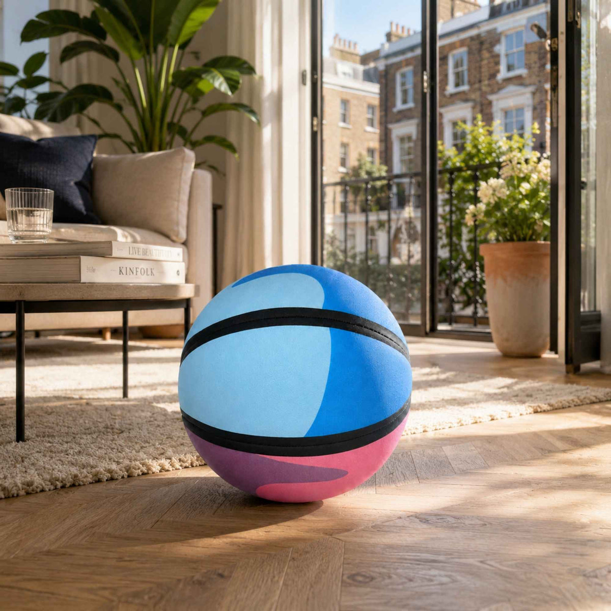 A HouseHoops basketball - SH with blue, pink, and black panels sits on a wooden floor in a bright, modern living room with large windows and a sofa in the background.