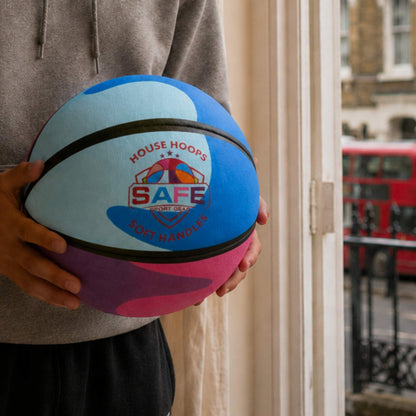 A person holds a HouseHoops basketball - SH in front of a window. The ball has a blue, pink, and light blue color-blocked design with black seams and a central logo that reads HOUSE HOOPS SAFE SPORT GEAR.