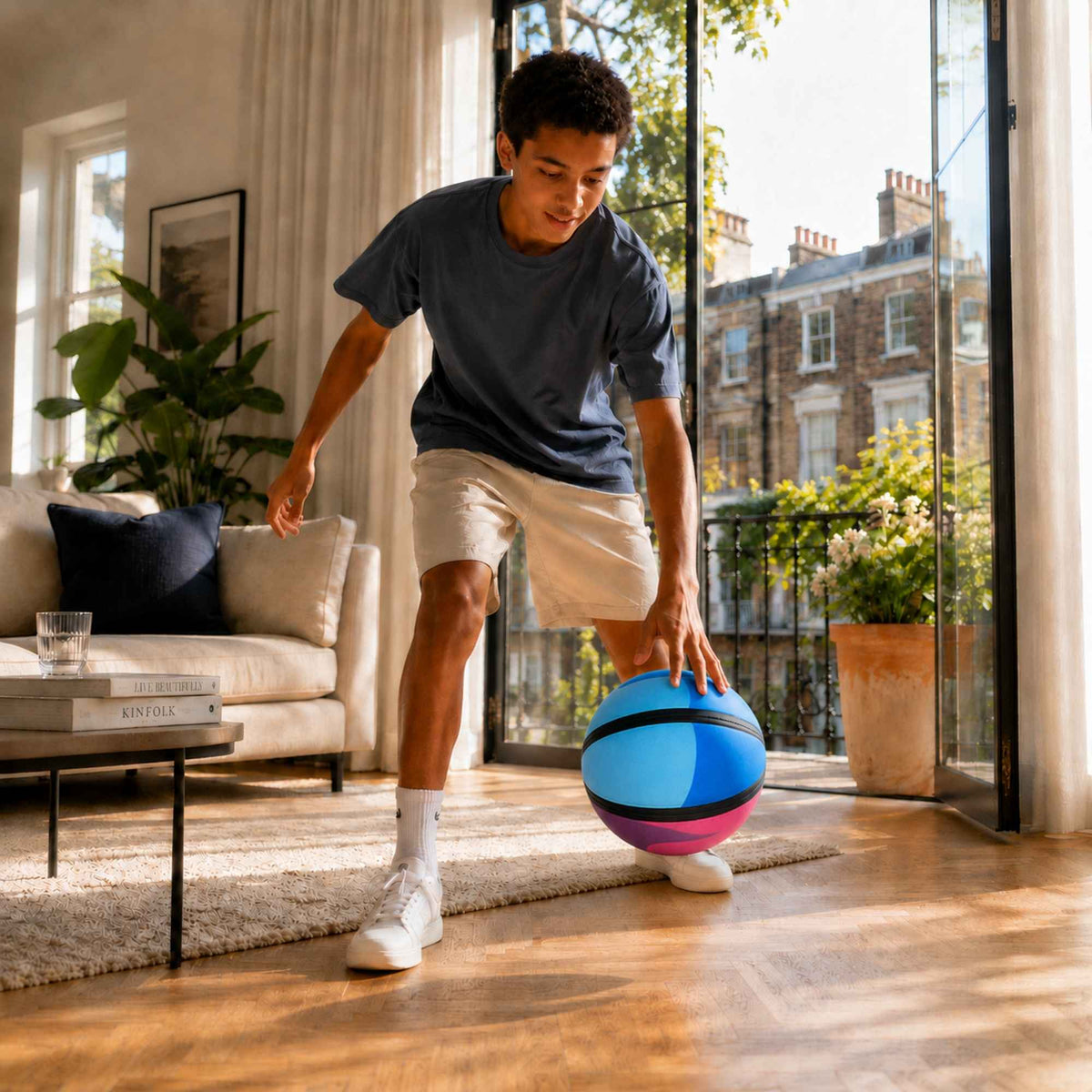 A man in a navy blue t-shirt and beige shorts dribbling a colorful HouseHoops basketball - SH in a bright, modern living room with large windows overlooking a city view.