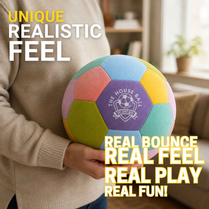A person in a beige sweater holds a colorful plush soccer ball with pastel panels and a purple center logo reading 'The House Ball'. The background is a softly blurred living room, with promotional text overlays reading 'Unique Realistic Feel' and 'Real Bounce Real Feel Real Play Real Fun!'.