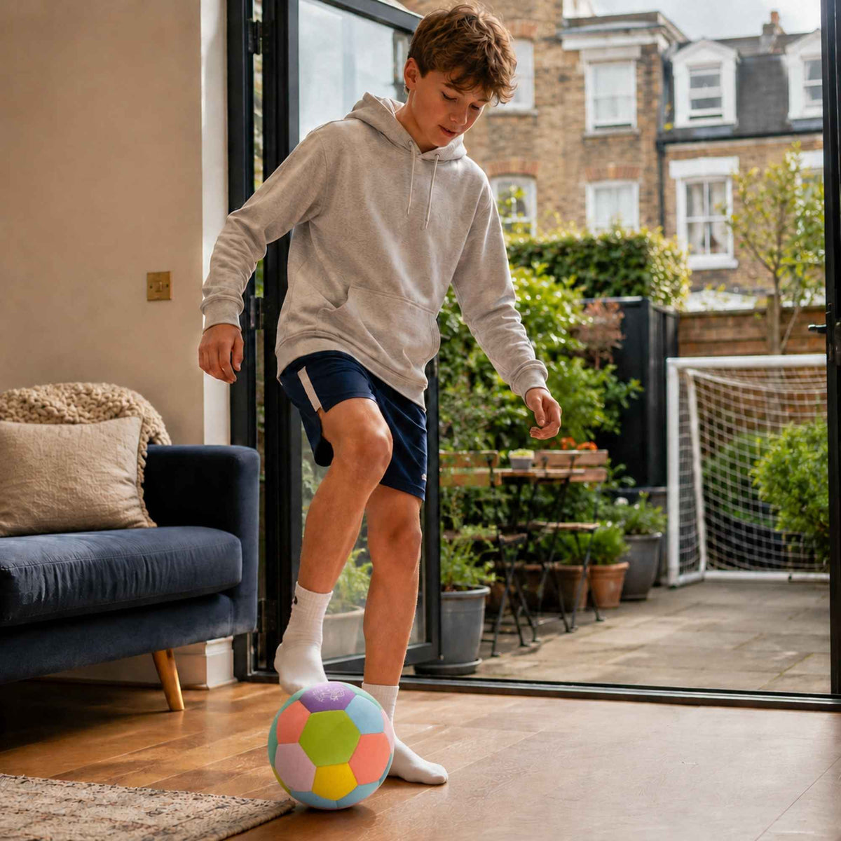 A young boy wearing a light gray hoodie, navy athletic shorts, and white socks is balancing a House Ball - EE on the wooden floor inside a modern living room, with a balcony and garden visible behind him.