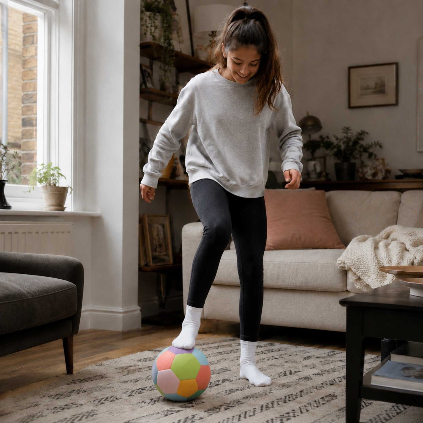A woman in a gray sweatshirt and black leggings balances a House Ball - EE on her foot in a cozy, sunlit living room.