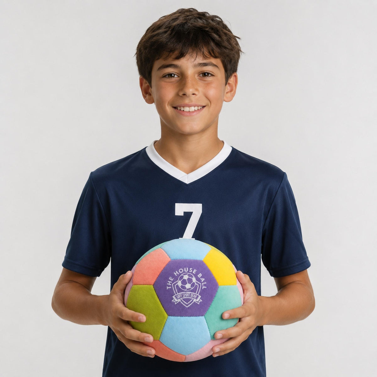 A smiling young boy with dark hair stands against a light gray background, wearing a navy blue soccer jersey with the number 7 and holding a colorful pastel House Ball - EE with a purple 'Safe Sport Gear' logo.