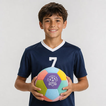 A smiling young boy with dark hair stands against a light gray background, wearing a navy blue soccer jersey with the number 7 and holding a colorful pastel House Ball - EE with a purple 'Safe Sport Gear' logo.